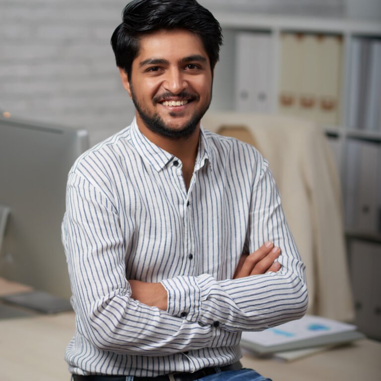Confident young Indian businessman sitting on edge of his table and looking at camera